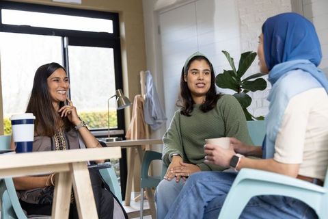 Multicultural women colleagues chatting over coffee in relaxed office lounge with greenery