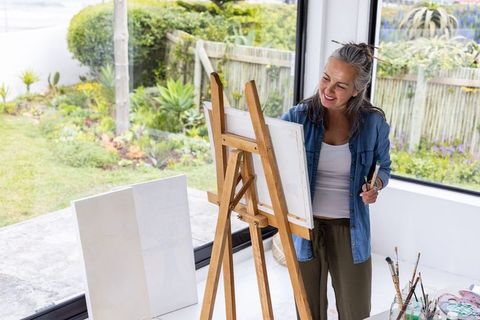 Senior artist painting at easel in sunlit home studio overlooking lush garden with brushes