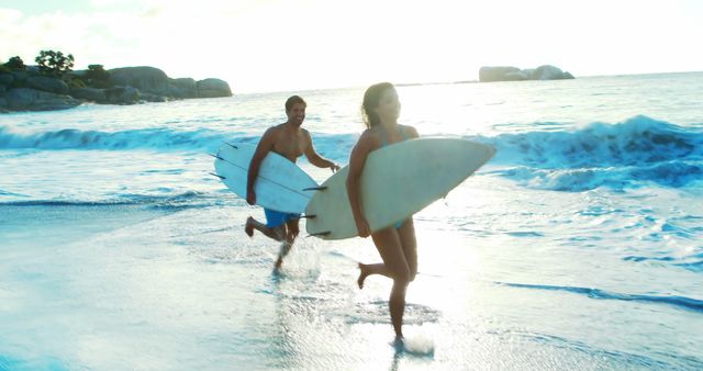 A couple is running through shallow water on the beach, holding surfboards, and smiling. The sun is setting, casting a warm glow on the scene, creating a romantic and energetic atmosphere. This image is perfect for promoting travel destinations, beach resorts, surfing events, outdoor activities, or fitness and lifestyle brands.