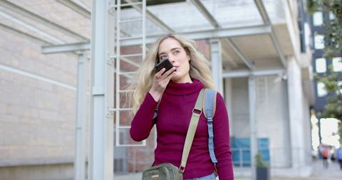 Young woman speaking into smartphone while walking with backpack and crossbody bag in city