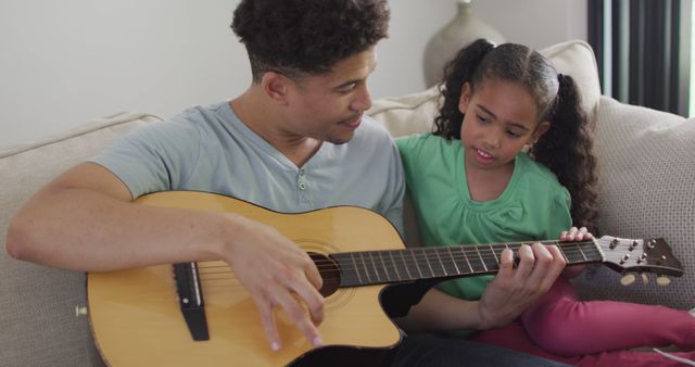 Father teaching young daughter to play acoustic guitar at home. Ideal for use in educational materials, parenting resources, family music enterprises, and lifestyle blogs focused on bonding and family activities.