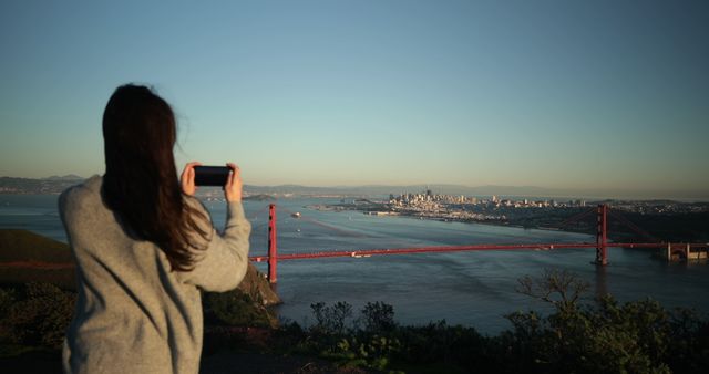 Rear view of a yong Caucasian woman with long dark hair standing in the Golden Gate National Recreation Area taking pictures of the Golden Gate Bridge and the San Francisco Bay Area with a smartphone 
