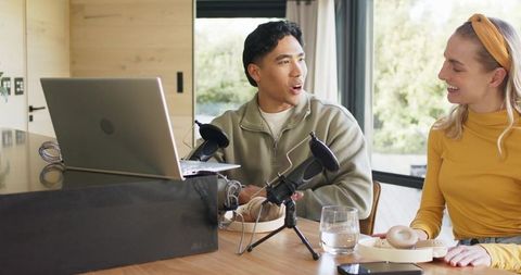 Diverse co-hosts recording home podcast at wooden table with laptop and microphones