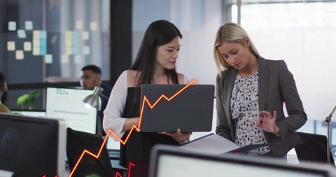 Two women collaborating on laptop and binder with upward sales graph in open office