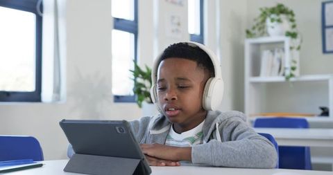 Student wearing white headphones using tablet for remote learning in bright classroom