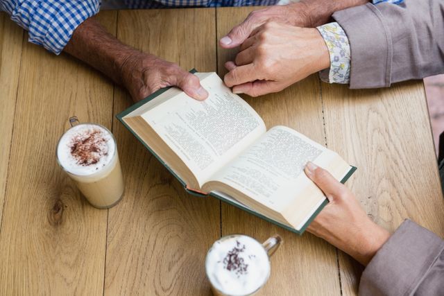 Senior couple enjoying a book together in an outdoor café, with two lattes on a wooden table. Ideal for use in articles or advertisements about senior lifestyle, retirement activities, bonding, and leisure time. Perfect for illustrating themes of love, friendship, and relaxation in later life.