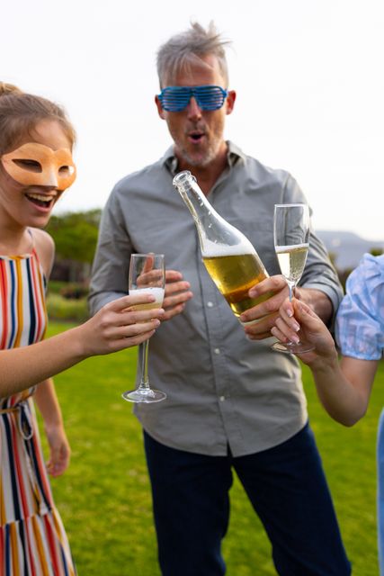 Man wearing party glasses pouring champagne into glasses held by two female friends in a garden. One woman is wearing a mask, adding a festive touch. Ideal for use in content related to celebrations, parties, social gatherings, and outdoor events. Perfect for illustrating themes of friendship, fun, and enjoyment.