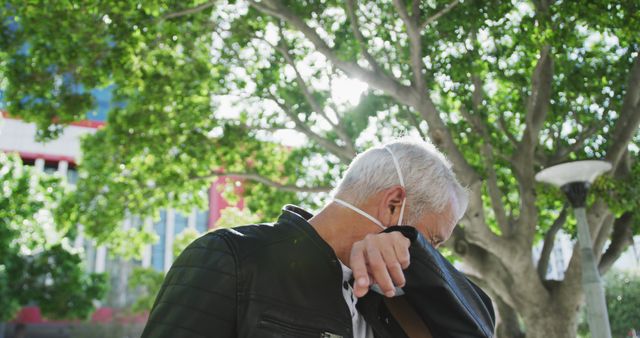 Mature man covering his cough with his elbow while outdoors under a tree. Ideal image for articles or campaigns focused on public health and safety, promoting hygiene, or raising awareness about respiratory protection in urban settings. Suitable for websites, blogs, or brochures on health and wellness.