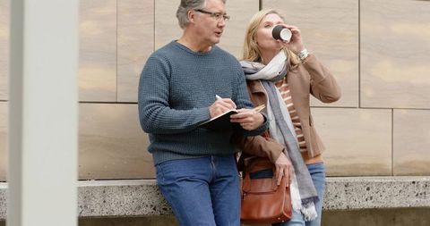 Senior couple leaning on bench in urban plaza man writing in notebook woman sipping coffee
