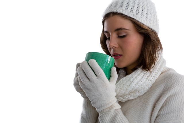 Woman dressed in warm winter clothing, including a white hat, scarf, and gloves, holding a green mug and enjoying a hot beverage. Ideal for use in advertisements for winter apparel, coffee or tea brands, and lifestyle blogs focusing on winter comfort and relaxation.