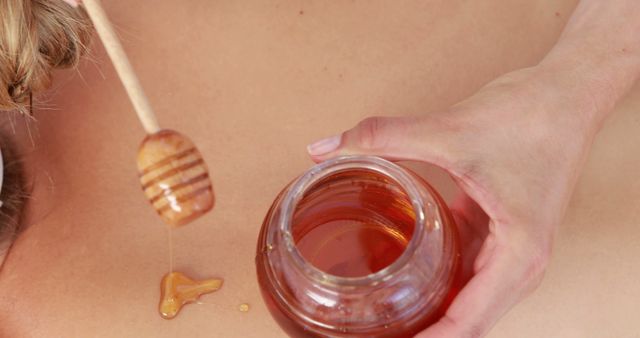 Close-up of a person's hand holding a jar of honey and using a wooden honey dipper to apply honey on the skin. The honey is dripping from the dipper, indicating a wellness or beauty treatment. This image is ideal for use in health and beauty products, spa promotions, natural therapies, or skincare brands. It can also be used in content about natural remedies or DIY beauty treatments.