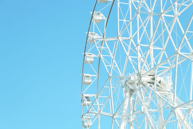 This image of a white Ferris wheel against a clear blue sky conveys a sense of fun and relaxation. The bright, cloudless backdrop suggests a sunny day, ideal for outdoor activities. This image can be used in travel brochures, leisure and tourism magazines, or advertisements for amusement parks and summer events.