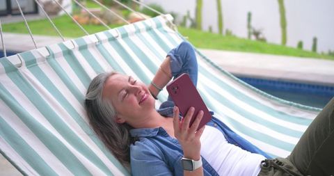 Senior woman reclining in striped hammock by pool relaxing and using smartphone
