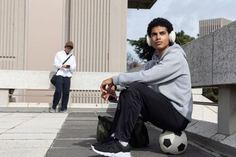 Diverse student wearing headphones sitting on soccer ball on urban rooftop terrace