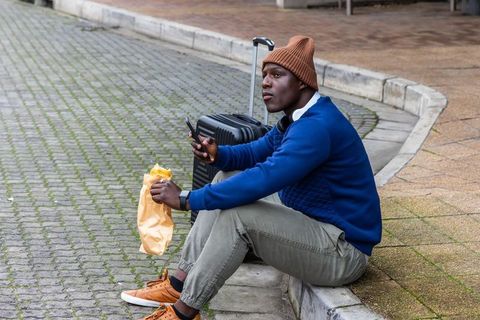 African american traveler sitting on curb eating sandwich checking phone with suitcase