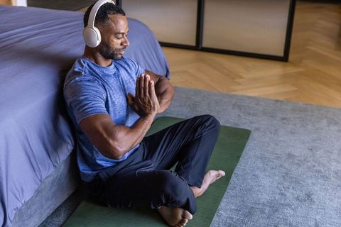 African american man meditating with headphones on yoga mat beside bed for mindfulness