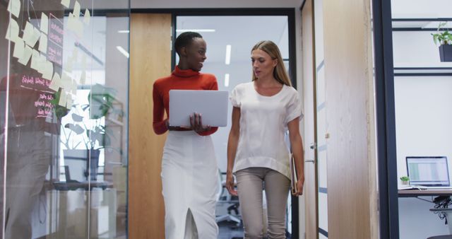 This image features two professional businesswomen engaged in a conversation while walking through an office hallway. One woman is holding a laptop and wearing a red turtleneck and white skirt, while the other woman, wearing light professional attire, is carrying documents. The setting is a modern office with open glass walls and visible workstations. This image can be used to represent themes such as teamwork, professionalism, corporate culture, diversity in the workplace, and collaborative work environments.