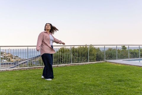Woman standing and turning on oceanview lawn terrace wearing pink jacket and white shoes