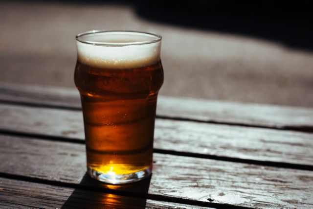 Chilled glass of beer on wooden picnic table outside in sunny environment. Ideal for use in food and beverage advertisements, outdoor leisure activities, summer events promotions, and articles about brewery craft beers.