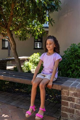 Young girl sitting on bench in shaded courtyard wearing lavender shirt and pink sandals