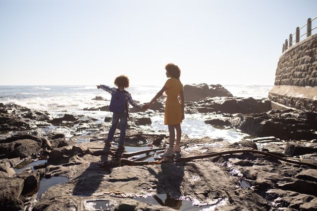 Mother and son enjoying a sunny day by the sea, standing on rocky beach and holding hands. The boy is pointing into the distance, suggesting exploration and adventure. Ideal for use in family, travel, and outdoor lifestyle themes, as well as advertisements promoting family bonding, vacations, and nature activities.