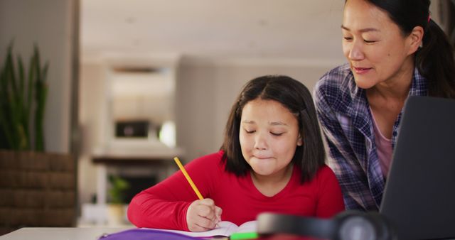 Mother assisting her daughter with homework in a cozy home environment. Ideal for use in education articles, parenting blogs, and family-related content. Illustrates supportive parenting, home schooling, and children learning.