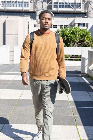 African american man walking with skateboard and headphones on urban campus in sunlight