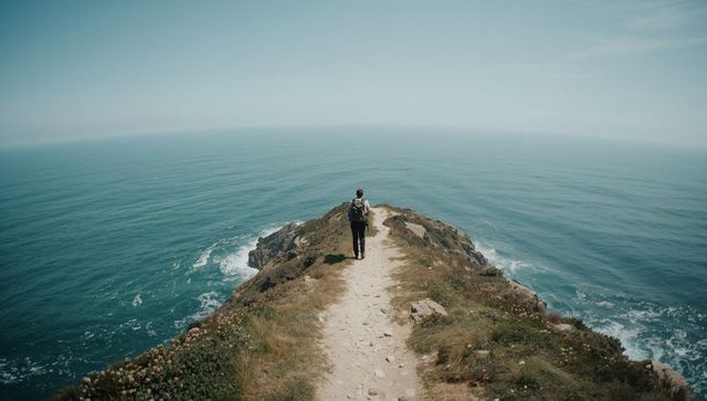 Solitary hiker walking narrow coastal trail toward cliff overlooking blue ocean horizon