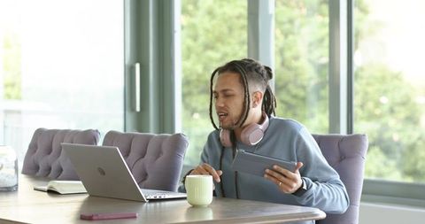 Mid adult African American man working from home with laptop and tablet reaching for mug