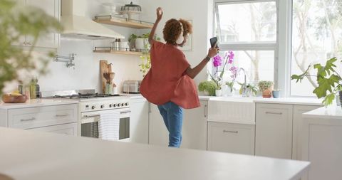 Woman dancing in modern kitchen wearing headphones holding smartphone near farmhouse sink