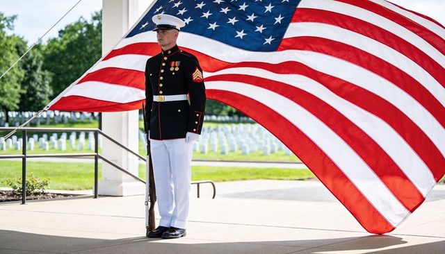 Marine standing guard beneath billowing american flag at national cemetery pavilion