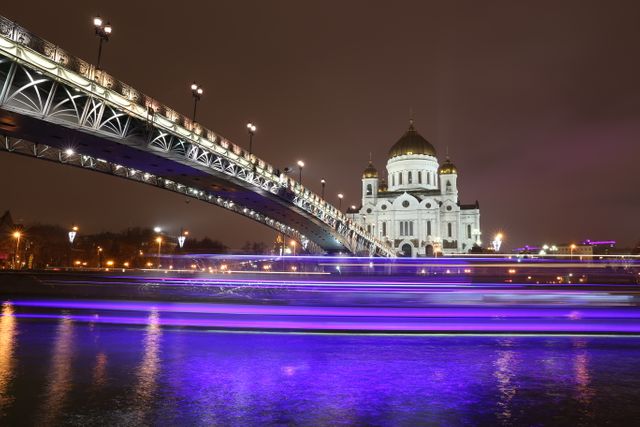 Capturing the majestic lighting of the cathedral and a bridge over the Moscow River during night time. Bright purple reflections shine on the water, giving the scene a modern touch. Perfect for use in travel guides, architectural illustrations, tourism websites, or cityscape presentations.