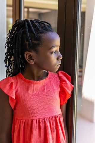 Contemplative african girl looking out sunlit window wearing coral dress