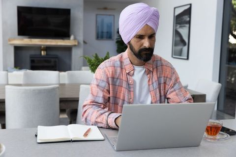 Mid-Adult Man in Turban Working at Home on Laptop with Notebook and Tea