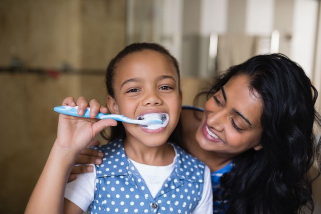 This image shows a mother encouraging her daughter to brush her teeth in a bathroom. The daughter is smiling while brushing her teeth, and the mother is looking at her with affection. This photo can be used for promoting dental hygiene, parenting tips, family health, and morning routines. It is ideal for websites, blogs, and advertisements related to dental care, parenting, and family well-being.