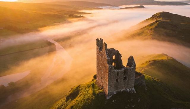 Ruined castle tower sitting on foggy hill at sunrise over winding river