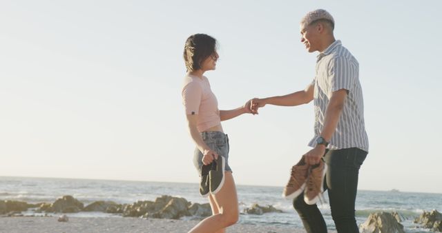 This image showcases a young couple enjoying a joyful moment together, holding hands and dancing barefoot on the beach. The carefree and sunny atmosphere highlights themes of love, romance, and happiness. This can be used for advertisements, travel blogs, romantic getaway promotions, social media content, or lifestyle websites focusing on relationships and leisure activities.