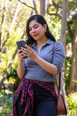 Young woman texting on smartphone while walking in sunlit park wearing casual outfit