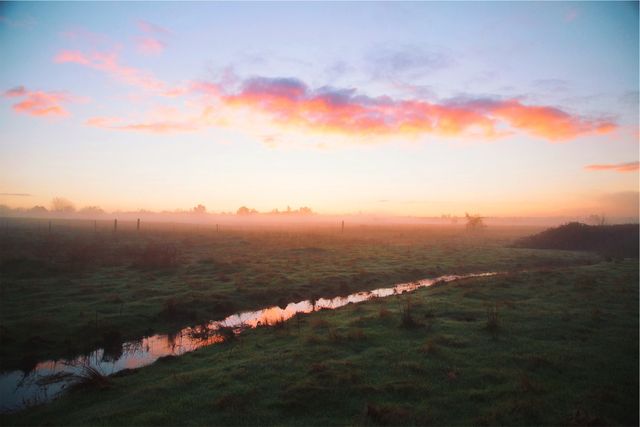 A peaceful morning landscape captures the first light of dawn, with warm colors spreading across the sky and reflected in a narrow stream running through misty fields. Ideal for nature-themed projects, relaxation settings, backgrounds, and promoting a calm atmosphere.