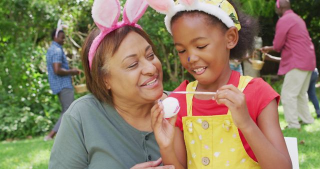 African American grandmother and granddaughter enjoy painting Easter eggs outdoors in a lush garden. Granddaughter dressed in yellow overalls with bunny ears carefully decorates an egg while grandmother looks on with a smile. Family members in background engage in festive activities. Ideal for use in content about family bonds, Easter celebrations, spring activities, cultural traditions, and multi-generational family moments.