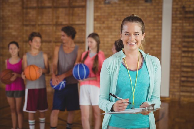 Smiling female coach holding clipboard in basketball court with young students holding basketballs in the background. Ideal for content related to sports education, youth training programs, coaching tips, fitness instruction, and team-building activities.
