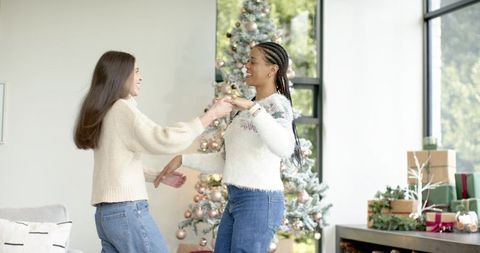 Two friends dancing by christmas tree in cozy sweaters during festive home celebration
