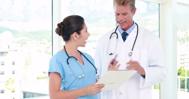 Medical professionals, a doctor and a nurse, are discussing patient data in a modern, sunlit clinic environment. The doctor is holding a clipboard, both are wearing uniforms and stethoscopes, illustrating teamwork and effective communication in a healthcare setting. Useful for depicting medical consultations, teamwork in healthcare, modern clinic environments, and healthcare facilities in promotional materials, informative articles, and educational content.