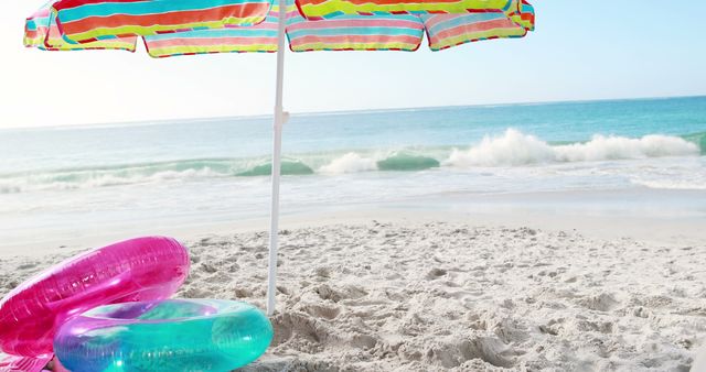Colorful beach umbrella and inflatable rings on sandy beach shore with ocean waves in background. Ideal for promoting travel destinations, vacation packages, or summer products. Perfect for use in magazines, tourism websites, and advertisements targeting beach lovers and summer vacationers.