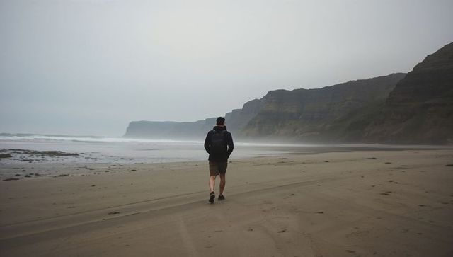 Solitary hiker walking misty coastline with backpack on overcast remote beach