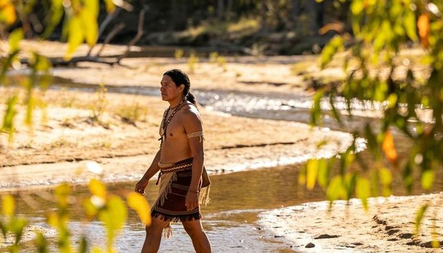 Indigenous man walking riverbank wearing traditional fringed wrap and beaded necklaces