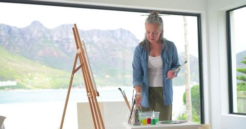 Mature woman painting in bright waterfront studio with easel, palette and mountain view