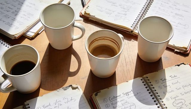 Sunlit table with five ceramic coffee mugs sitting among handwritten notebooks and shadows