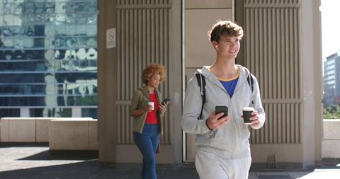 Young commuters walking holding takeaway coffee and checking phones in urban plaza