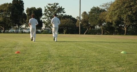 Diverse male cricketers walking across grassy field in white kits passing training cones